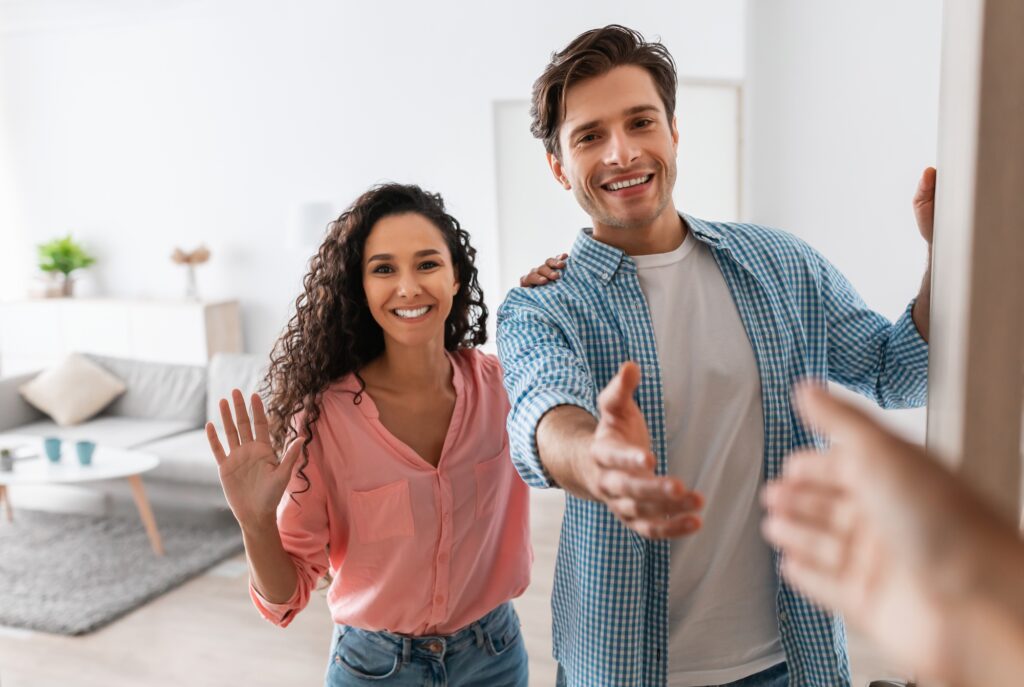 Happy Young Couple Inviting People To Enter Home Shaking Hands