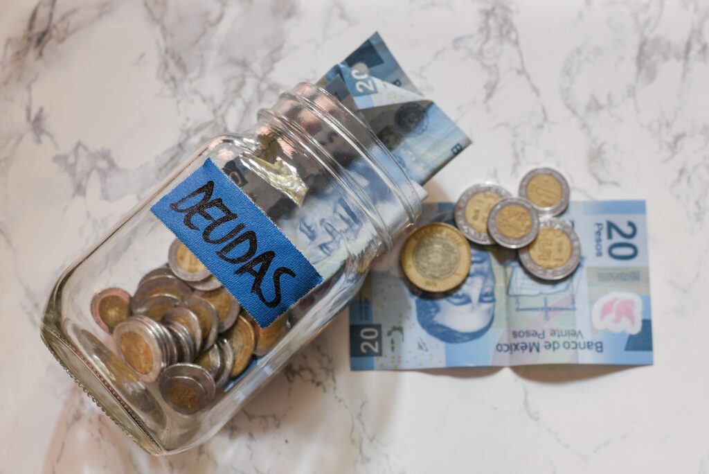 High Angle View Of Pesos And Coins In A Jar With A Blue Deudas Debts Sticker On It On The Table